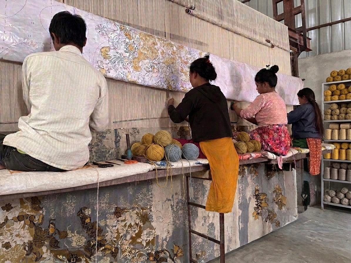 Several weavers working beside a large loom in a decorative rugs workshop in Kathmandu, weaving rug with visible yarn skeins and a partially finished. Shelves with yarn are visible on the right.